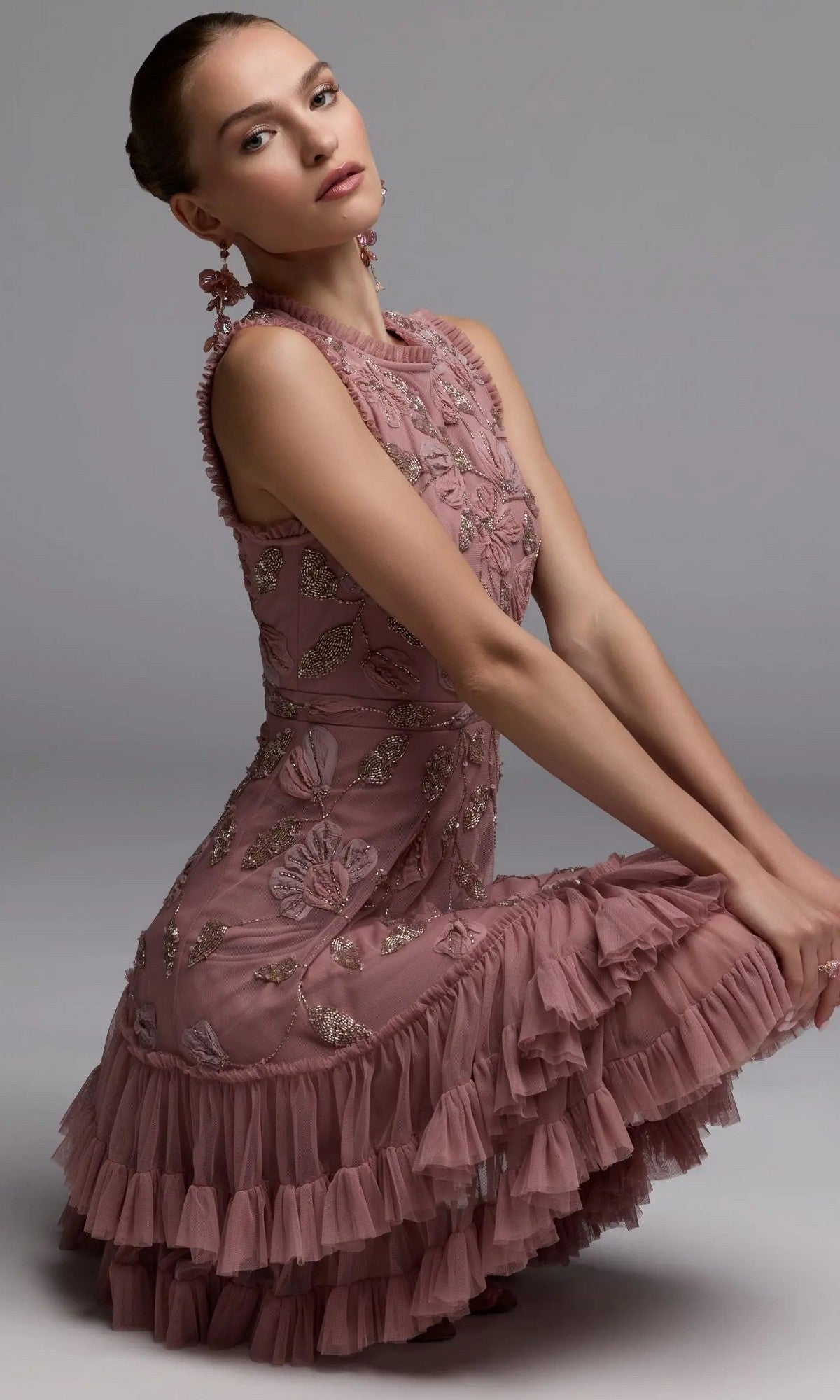 A woman kneels, looking at the camera, wearing the Mac Duggal 9403 Short Party Dress—a mauve fit-and-flare style with ruffles and floral embroidery—against a plain gray background.